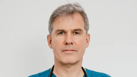 Dr Rod Mackenzie smiles at the camera. He has short grey hair and stands against a white wall. He wears a blue doctor scrubs as well as a black stethoscope around his neck