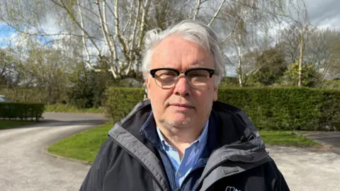 A man wearing glasses and a black raincoat is standing outdoors. He has grey hair and stubble around his chin. He is looking directly at the camera.