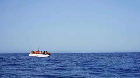 A vast open blue ocean and sky with a small white boat packed with people in orange lifevests