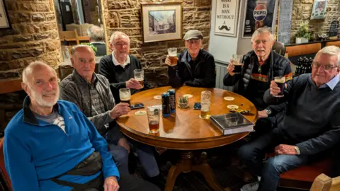 Six gentlemen in their eighties sit around a pub table holding their beers up.
