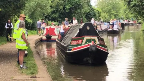 Martin Heath/BBC Canal scene, showing several narrowboats. The boat nearest the camera is painted in red and green and has the word KIMBERLEY in white lettering on the side. There is a canopy at the front of the boat. There are three people at the back. There are two people wearing hi-vis vests in the foreground. There are many other people and boats in the background.