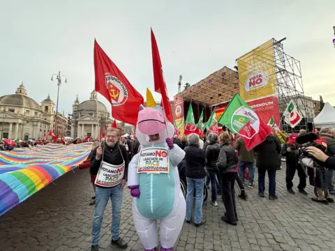 A crowd of people carry flags with the hammer and sickle. There is a large rainbow flag held above the ground. The protestor closest to the camera is dressed in an inflatable unicorn costume. 