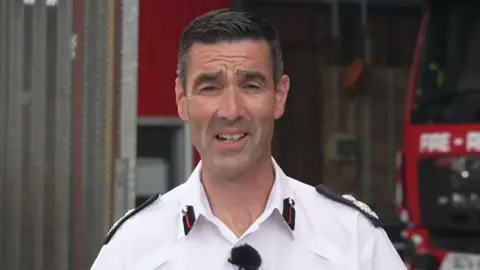 The Devon and Somerset fire's services's assistant chief is wearing a white shirt with black epaulettes. He is standing in front of a red fire engine.