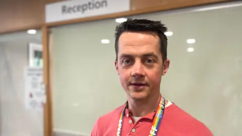 Mark Norman BBC Mikey Pearce, a  Youth Emotional Support Worker looks at the camera in front of a GP reception desk