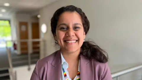 A woman stands at the top of a staircase in a bright hallway at a University building. She is wearing a pink jacket and a rainbow lanyard. 