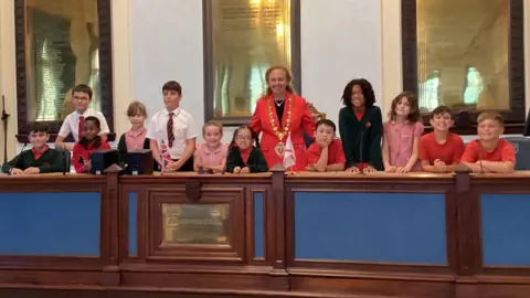 12 children in school uniform and a woman with a big gold ceremonial chain around her neck a red blazer on. They are standing on a stage in the town hall, leaning on a table and behind them big plaques with inscriptions on.