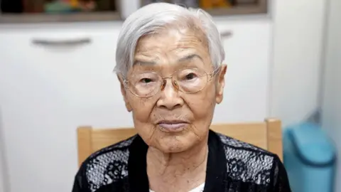 BBC/HYOJUNG KIM Uma velha mulher branca branca e os óculos de arame está em uma cadeira e olha para a câmera