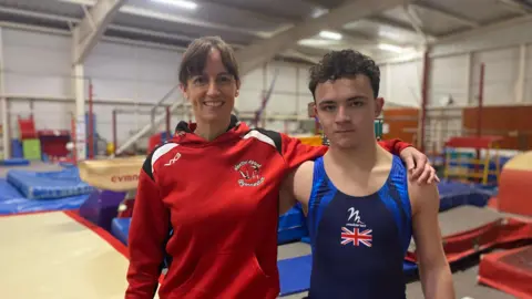 A man standing inside a gymnasium wearing a UK navy blue leotard. A woman wearing a red hoodie has her arm around him.