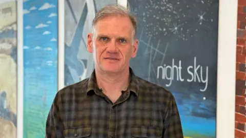 Dr Steve Wallace wearing a brown chequed shirt standing in front of displays at the observatory