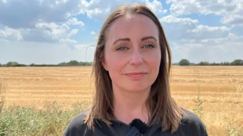 A woman standing in front of a large arable brown field on a summery day. There are white clouds above.