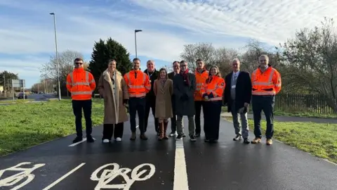 North Northamptonshire Council A group of people, some in orange high-visibility jackets standing on the cycle/pedestrian path, which has a thick white line down the middle, with the left had side divided into two lanes for cycles - one for each direction of travel. 