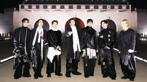 k-pop group BTS pose for aphotograph. They are all standing in a line wearing black with some white accessories and holding microphones