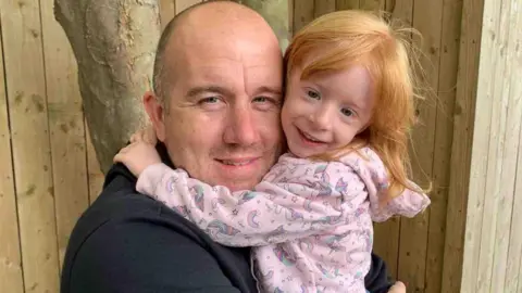 Handout Mark Prince, who has a bald head and is wearing a black hoodie, holds his daughter Holly who has ginger hair and is wearing a pink unicorn themed jumper. Both are smiling at the camera