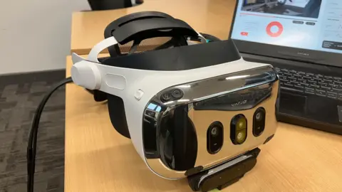 A VR headset in white and black, on a desk in front of a computer screen 