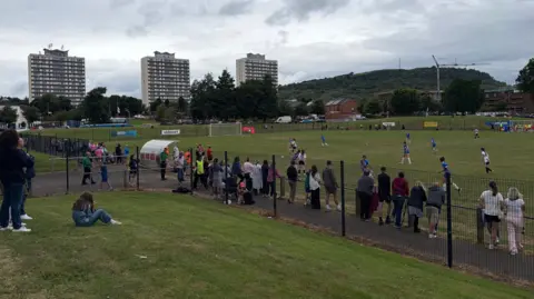 The Diamond in Rathcoole, a football pitch surrounded by a black fence. Parents watch the game between Crusaders in white and Surf Select in Blue. Three high-rise blocks of flats are to the left behind the pitch.