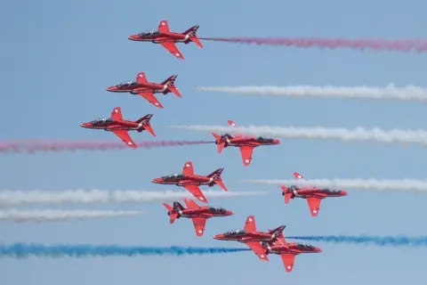 David May There are nine Red Arrows in the picture, some are pointing in different directions. There are red, white and blue smoke trails.