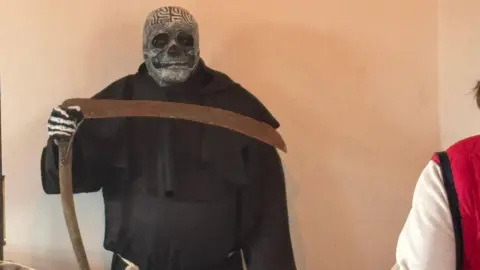 Louise Miller A man dressed as the Grim Reaper standing at the back of a funeral