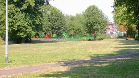 A small park in Northampton, showing green grass, a part of a lamp post to the left, a playground, in the distance, with green and red apparatus and trees. Buildings are to the right. 