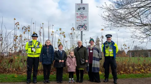 Office of Northamptonshire's Police, Fire and Crime Commissioner Two police officers in hi-vis jackets stand next to three children, a woman in an olive coat with glasses, and a woman in a grey, navy and grey striped top. They are standing in front of a metal pole with the anti-nuisance motorbike signs