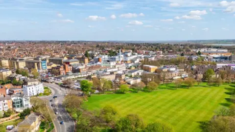 An aerial view of a Northampton on a clear day. The scene shows the greenery of Beckets Park, beside the busy Bedford Road junction. In the background is the expansive Northampton General Hospital site. The height of the aerial means that the view to the horizon extends for several miles across rooftops and fields.