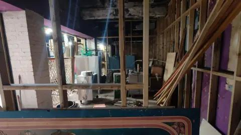 Inside the empty C103 building. The room is run down with wooden structures exposed. Purple wall paper is deteriorating. The space is filled with miscellaneous items. 