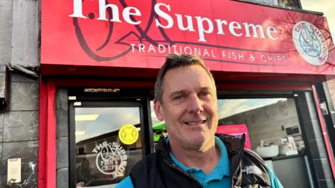 Neill Edgar. He has short brown hair, wearing a black North Face gilet and blue polo top. A red shop front for The Supreme traditional fish and chips is behind him. 