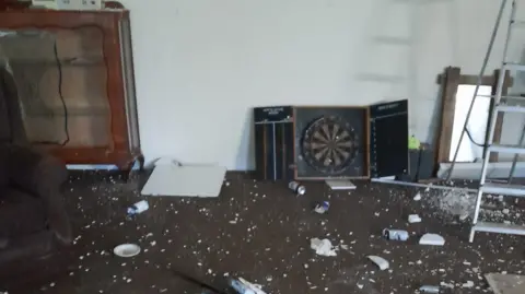 Stone Town FC Debris strewn across the floor of a clubhouse. A dartboard is on the floor against the wall. A ladder can be seen on the right and a cupboard on the left.