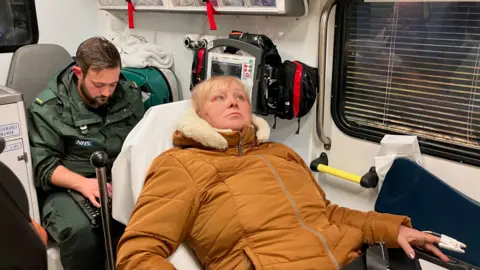 Sarmite Sinkeviča, wearing an orange overcoat, lies on a stretcher in an ambulance with paramedic Shane Guinan sitting behind her working on a laptop.