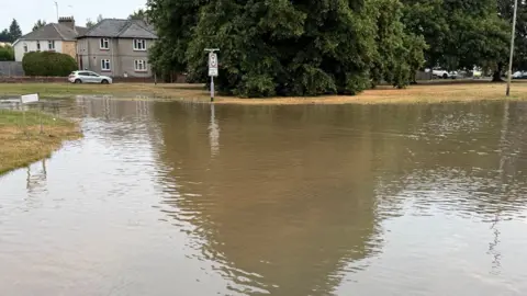 A road, where a junction would be, has flooded and the water has gone above pavement level. There are houses and trees in the background and there are several cars.