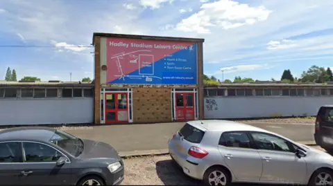 Google A low building, which looks slightly run-down, next to a road where cars are parked. In the middle of the building is an entrance above which is a sign reading "Hadley Stadium Leisure Centre".