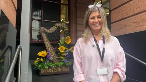 A woman with blonde hair smiling outside Central Library, a red brick building. She is wearing a pink blouse nad in the background is a large wooden harp sculpture with bright yellow flowers on it. It reads Belfast Fleadh Cheoil.