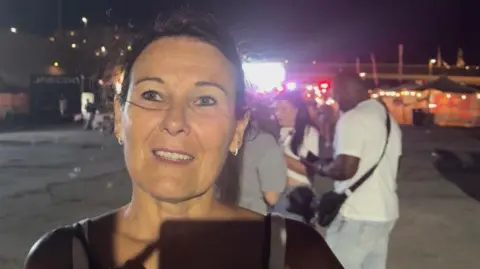 BBC / Robin Grey Elaine Connolly stands outdoors at night speaking to camera, with bright lights, stalls and several people visible in the background.