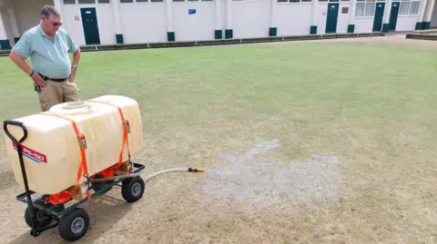 Steve O'Dare A man in a blue polo shirt watches as a small water tank empties water through a hose on to a dry-looking bowling green.