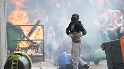 Getty Images An image of rioting in Rotherham in August 2024. A protestor stands in the foreground of the image wearing grey tracksuit bottoms and a covering over his face and head. A crowd is in the background, behind a bin on fire next to a Holiday Inn Express