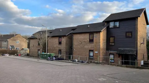 A general view of low-rise flats with bin bags piled in front in one area and paved parking. Police tape can be seen tied to a fence in the left of the photo. 