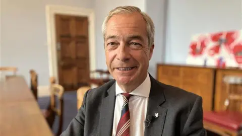 Close up image of Nigel Farage wearing a suit sat in a council chamber at County Hall in Maidstone