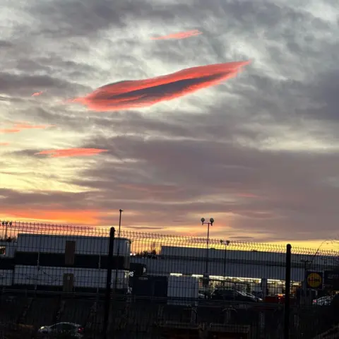 Colmc/BBC Weather Watchers The cloud glows an orange-pink above industrial buildings that are behind a tall wire fence.