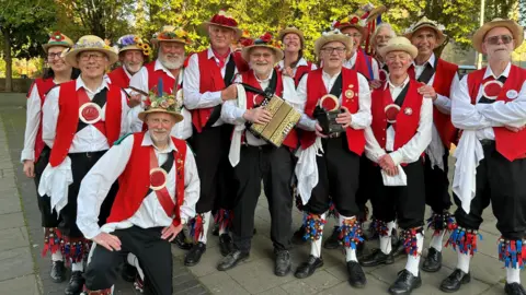 Joe Lemer/BBC A group of male morris dancers stand together as they face the camera. They are all wearing white shirts and red waistcosts with colourful tassels and bells around their shins. The men, morris dancers from Bristol, also all have straw boater-style hats on
