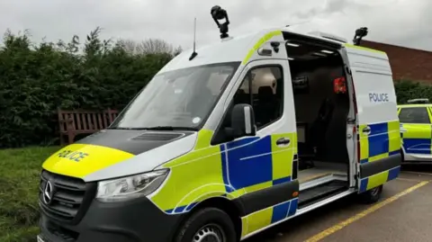 Jack Fiehn/BBC A white, yellow and blue police van parked by the side of the road, with the side door open