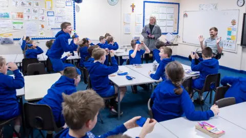 Everton School children look happy and surprised as they see the footballer in their classroom. Kiernan smiles and holds his hands up at the front of the room.