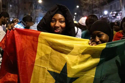NurPhoto via Getty Images Fãs de futebol senegaleses comemoram em Paris. Duas mulheres seguram a bandeira do Senegal.