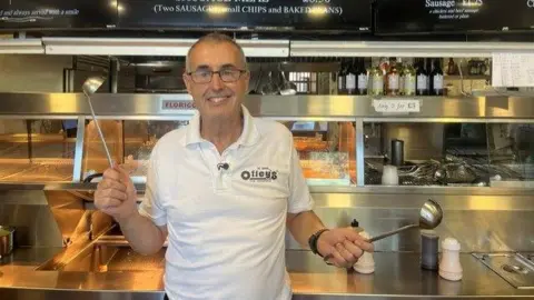 A middle aged man with glasses wearing a white polo shirt stands in a restaurant kitchen. He is hold ladels in his hands.
