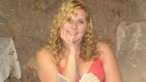 PA Media Melissa Power sits holding an open book and and looks at the camera smiling. She has long blonde curly hair and is wearing a coral-coloured skimpy top. She is inside a building with rough stone walls and the corner of a table with a table cloth is to the left of the picture.