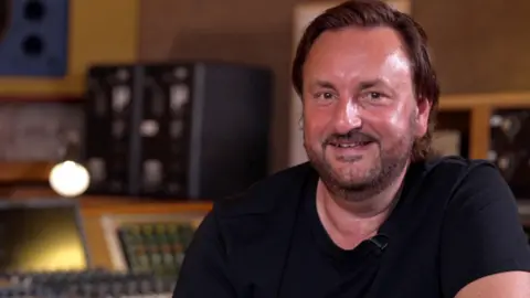 Nick Brine now. He has chin-length dark hair, slicked back, and is wearing a black t-shirt with the recording studio desks blurred in the background.