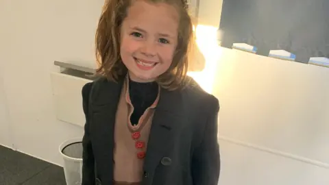 Stagecoach Garforth A little girl smiles into the camera wearing a grey blazer and a brown jumper and red buttons.