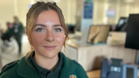 Hannah Dickson is pictured wearing her dark green ambulance service uniform. She has long brown hair tied back from her face. 