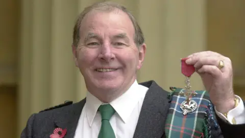 PA Media Sydney Devine smiles at the camera. He wears a kilt with a grey blazer, green tie and white shirt, with tartan wrapped around his shoulder and a red poppy pinned to his chest. He holds his MBE up for the camera.
