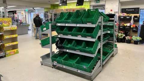 A large unit of green baskets which are all empty in the Co-op in St Mary's. There is an empty display headed EASTER and there are buckets of flowers.