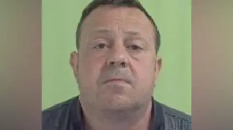 ISLE OF MAN CONSTABULARY Cannon's police mugshot. He has short dark hair and stubble and is wearing a dark grey T-shirt with a black collar.