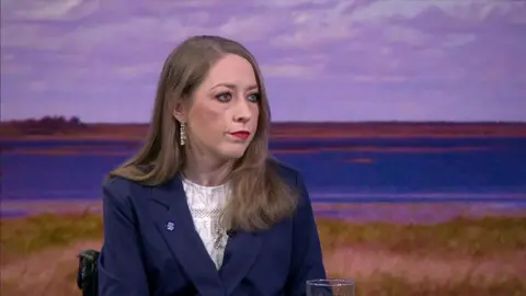 Dr Clodagh Corrigan is sitting down at a table in front of a backdrop depicting a lake. She has long brown hair. She is wearing a dark blue blazer, a white top and a small pin on her lapel.